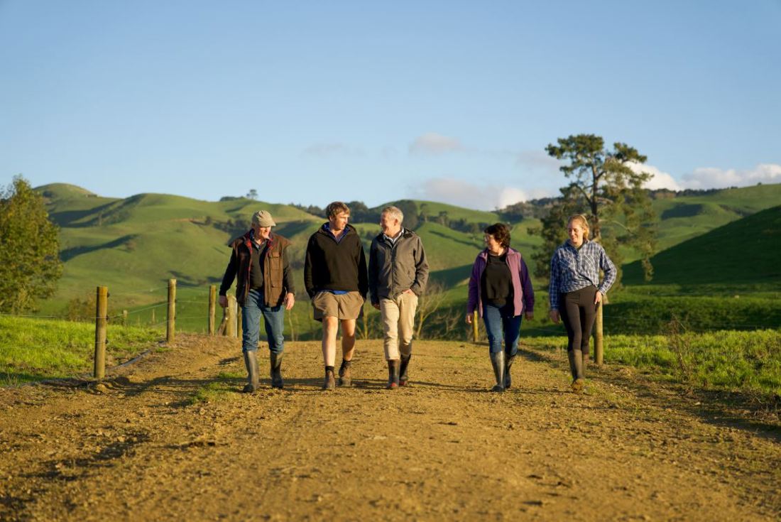 Five New Zealand dairy farmers walking along a farm track with green hills, trees and fencing in the background